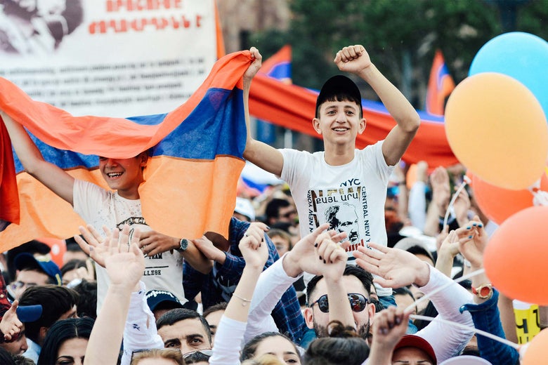 Young protesters ride on the shoulders of others amid a crowd carrying balloons and flags.